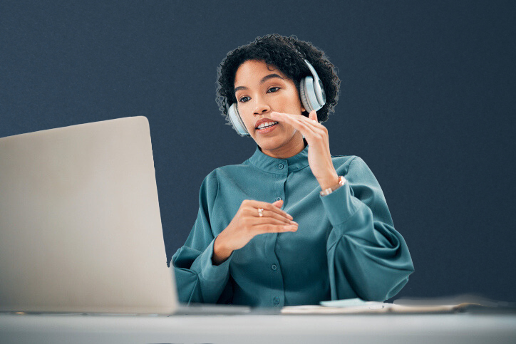 A woman with curly hair wearing a headset, gesturing with her hand as she participates in a video call on her laptop.