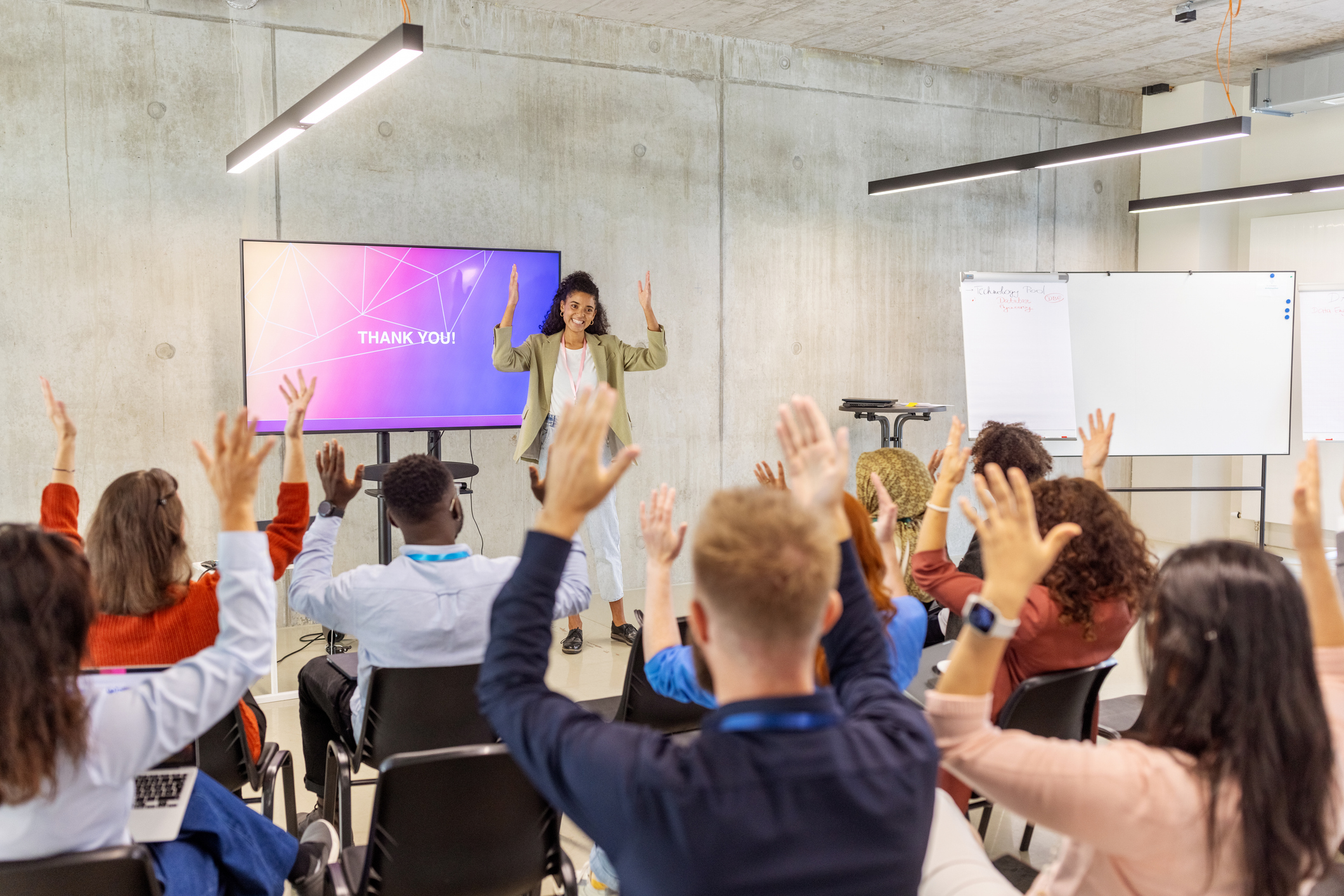 A woman stands on stage, signing applause in American Sign Language with enthusiasm as the screen behind her displays ‘Thank You!’, while a diverse audience of seated people applaud with their hands.