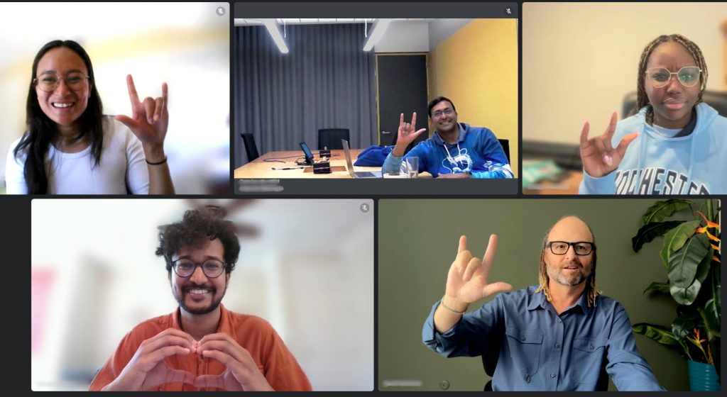 A screenshot of a video conference showing five people in individual frames, all using sign language. Some are making a hand gesture resembling ‘I love you’ while smiling.